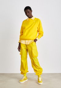 Felpa gialla con colletto sopra una maglietta più chiara, abbinata a pantaloni cargo e sneakers, tutto in giallo coordinato. Tessuto morbido, vestibilità rilassata.