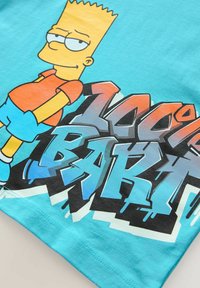 Tirkīzkrāsa kokvilnas t-krekls ar grafiku, kurā redzams karikatūras varonis oranžā kreklā un zilās biksēs, ar tekstu "100% BART" treknrakstā, stilizētās burtos.