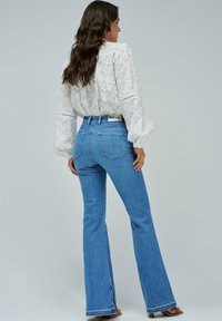 Los pantalones vaqueros acampanados de un azul claro cuentan con una cintura alta, diseño de cinco bolsillos y costuras sutiles. Se combinan con una blusa blanca texturizada que presenta bordados florales.