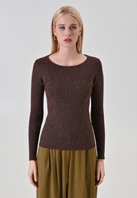 Jeune femme aux cheveux blonds raides, portant des boucles d'oreilles en or, un haut ajusté à manches longues marron en maille et un pantalon vert olive, se tenant devant un fond gris.