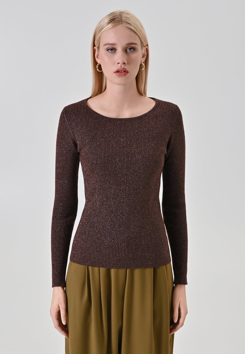 Jeune femme aux cheveux blonds raides, portant des boucles d'oreilles en or, un haut ajusté à manches longues marron en maille et un pantalon vert olive, se tenant devant un fond gris.