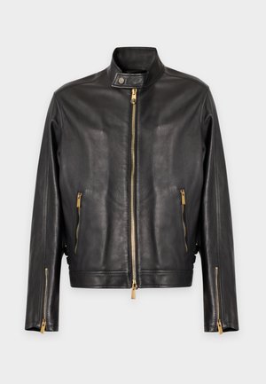 Jeune homme debout portant une veste en cuir noire, une chemise rayée, un cardigan blanc, un jean bleu, une ceinture noire et des mocassins noirs sur un fond uni.