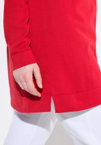 Maglioncino rosso lavorato a maglia con maniche lunghe, spacco laterale all'orlo e polsini a coste, abbinato a pantaloni bianchi. Texture morbida.