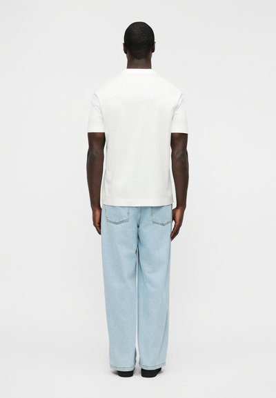 Άσπρο t-shirt με κοντά μανίκια, σε χαλαρή γραμμή, συνδυασμένο με ανοιχτόχρωμο τζιν παντελόνι με φαρδιά μπατζάκια, εμφανιζόμενο από πίσω, σε απλό φόντο.