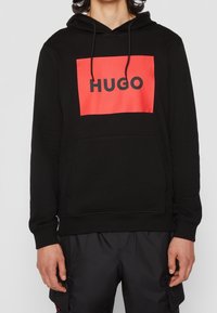 Felpa nera con cappuccio caratterizzata da un importante grafico rettangolare rosso con "HUGO" in bold nero. Il tessuto appare liscio e texture.