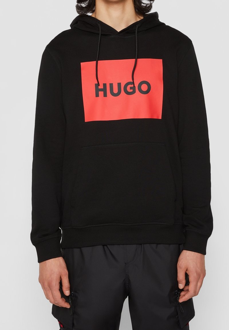 Felpa nera con cappuccio caratterizzata da un importante grafico rettangolare rosso con "HUGO" in bold nero. Il tessuto appare liscio e texture.