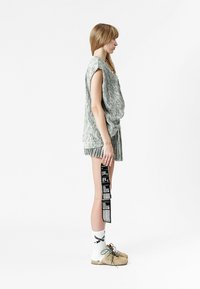 Top sin mangas de color gris claro y pantalones cortos plisados a juego, sosteniendo una etiqueta impresa. Los accesorios incluyen calcetines con un estampado gráfico y zapatos beige.