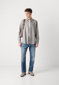 Grijze blouse met lange mouwen en knoopsluiting, blauwe denim jeans en lichtgrijze sneakers. De blouse heeft een logo op de voorkant en een relaxte pasvorm.