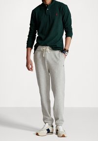 Grünes langärmliges Polohemd mit kleinem rotem Logo, graue Sweatpants mit elastischen Bündchen und beige Sneakers mit marinefarbenen Akzenten.