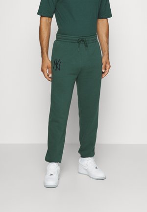 Pantalones de chándal verdes con cintura elástica y cordón, con un logo oscuro en el muslo izquierdo. Combinados con zapatillas blancas.