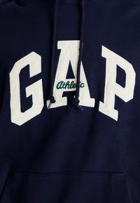Felpa blu navy in cotone, con grandi lettere crema "GAP" e testo verde "Athletic", dotata di tasca a marsupio frontale e cappuccio con cordino.