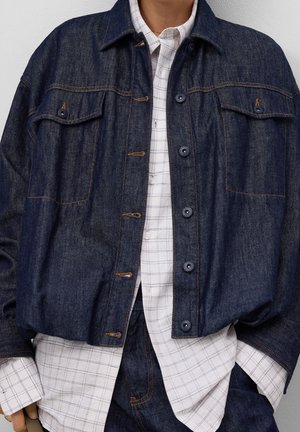 Person trägt eine dunkelblaue Jeansjacke über einem weißen karierten Hemd mit Knöpfen und passenden dunklen Jeanshosen, die Hände teilweise in den Taschen.