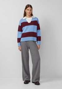 Strickpullover mit blauen und kastanienbraunen Streifen, weißem Kragen und lockerer Passform. Kombiniert mit grauen weit geschnittenen Hosen und Schuhen mit dicker Sohle.