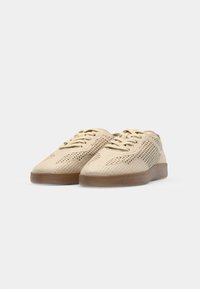 Paire de baskets beiges en tricot à lacets avec semelles en caoutchouc marron sur un fond clair uni.