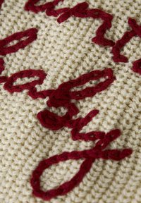 Tessuto di maglia color crema con testo ricamato in rosso scuro, dotato di un design testurizzato e a anelli. Le fibre hanno un aspetto morbido.