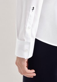 Camicia bianca a maniche lunghe, con polsini a bottone e un piccolo ricamo di rosa nera sulla manica. Tessuto dalla superficie liscia.