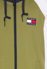 Oliwkowa kurtka z granatowym kapturem, z zamkiem z przodu i naszywką Tommy Jeans, która ma czarne, czerwone i białe paski. Gładka tekstura, swobodny design.