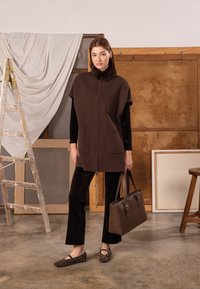 Brown zip-up vest with a collar and front pockets, paired with black velvet pants and studded brown flats. Holding a brown handbag.