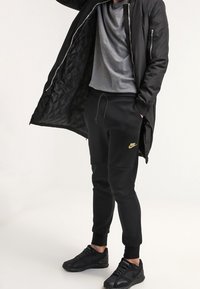 Black puffer jacket with quilted lining, gray t-shirt, black joggers with gold Nike logo, and black athletic shoes.