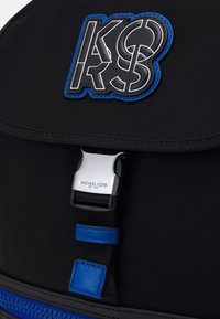 Sac à dos en nylon noir avec un patch logo bleu et noir, une fermeture à boucle en métal argenté et des accents bleus sur la fermeture éclair.