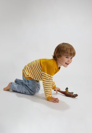 Pull rayé jaune et blanc, jean en denim bleu, enfant rampant sur le sol avec un dinosaure en jouet à la main, pieds nus, fond gris clair.