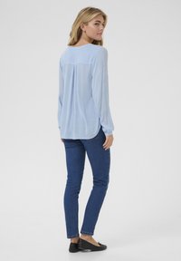 Blouse bleu clair à manches longues avec un col rond, coupe décontractée et détail de pli dans le dos, assortie à un jean bleu foncé ajusté et des ballerines noires.