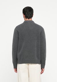 Uomo di spalle che indossa un maglione a maglia grigio scuro e pantaloni beige chiaro su uno sfondo bianco uniforme.