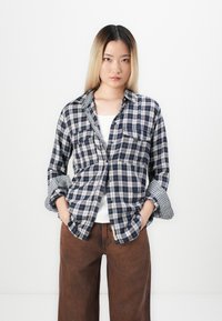 Plaid button-up shirt in navy and white, featuring two chest pockets and rolled-up sleeves. Paired with brown wide-leg trousers.