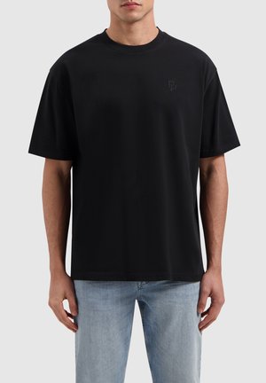 Black cotton T-shirt with a relaxed fit, crew neck, and short sleeves. Embroidered logo on the chest, smooth texture. Worn with light blue jeans.