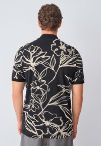 Juoda polo marškinėliai su dideliu smėlio spalvos gėlių raštu ir lapų motyvais. Trumpomis rankovėmis, plokščiu apykakle, pagaminti iš minkštos medžiagos, šiek tiek prigludę.