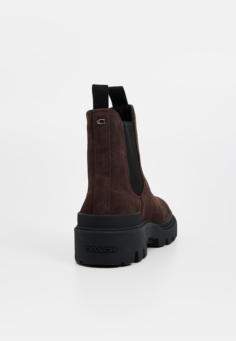 Bottines en suède marron avec des panneaux élastiques sur les côtés et des semelles en caoutchouc noires. Présente un logo à l'arrière et une semelle épaisse pour une bonne traction.