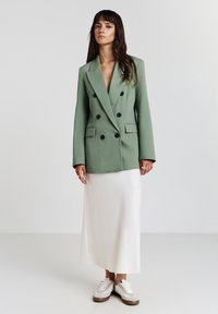 Blazer vert croisé avec des boutons noirs, associé à une jupe midi blanche et des baskets beiges. Tissu lisse avec une coupe structurée.