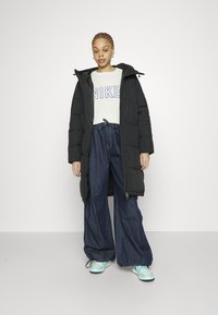 Zwarte pufferjas met een capuchon, oversized witte sweatshirt met blauwe letters, wijde spijkerbroek en lichtblauwe sneakers.