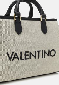 Vaaleanbeige teksturoidulla käsilaukulla, jossa on mustat nahkakoristeet ja -kahvat. Varustettu kultaisilla yksityiskohdilla ja "VALENTINO"-teksti suurilla mustilla kirjaimilla.