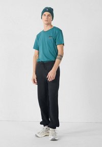 Teal T-shirt met korte mouwen en logo, gecombineerd met zwarte broek met trekkoord. Model draagt een donkergroene beanie en lichte sneakers. Effen achtergrond.