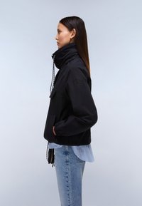 Mujer con chaqueta negra de cuello alto y manos en los bolsillos, camisa de rayas azul claro y jeans azules, de perfil contra un fondo liso.