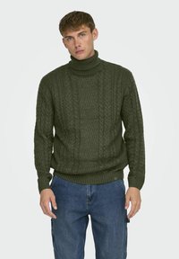 Suéter de punto verde oliva con cuello alto, que presenta un patrón de trenzado texturizado y puños y dobladillo acanalados, combinado con jeans azules.