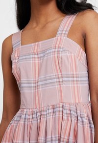 Jasnoróżowa sukienka w kratkę z kwadratowymi ramiączkami, plisowaną spódnicą i kontrastującymi paskami w odcieniach szarości i koralu. Bawełnowy materiał, casualowy design.