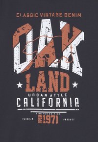 Svart t-shirt med fet, sliten vit och orange grafisk text som läser "OAK LAND URBAN STYLE CALIFORNIA" med stjärnaccent.