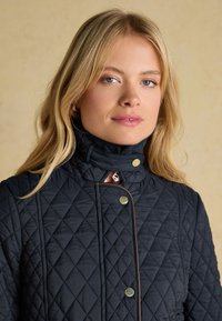 Blonde woman with blue eyes wearing a black quilted jacket with gold snap buttons and a high collar against a beige background.