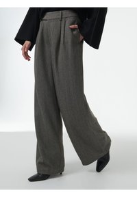 Persoon in een grijze herringbone broek met wijde pijpen en met de handen in de zakken, gekleed in zwarte, puntige hakken, staande op een witte achtergrond.