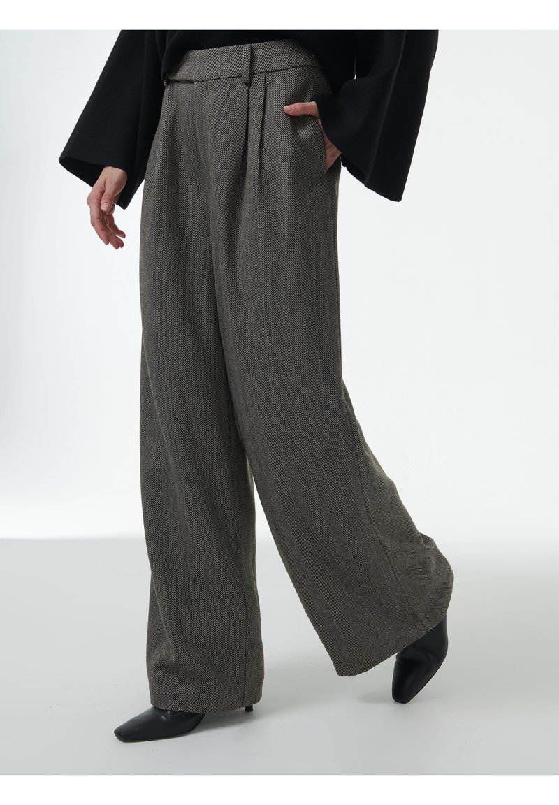 Persoon in een grijze herringbone broek met wijde pijpen en met de handen in de zakken, gekleed in zwarte, puntige hakken, staande op een witte achtergrond.