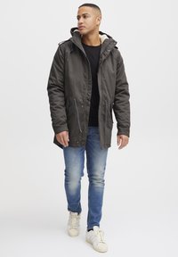 Parka de color verde oscuro con capucha forrada de piel, que cuenta con bolsillos con cremallera y cordones. Llevada sobre una camiseta negra y jeans azules.