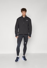Zwarte sportjack met hoge kraag en ritssluiting, gecombineerd met zwarte legging met zijaccenten en een logo, gedragen met marineblauwe hardloopschoenen.