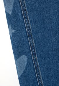 Denimstoff, dunkelblau mit helleren Sternmustern, mit kontrastierenden Nähten entlang der Naht, die die strukturierte Oberfläche betonen.