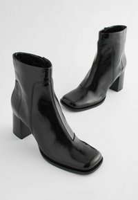 Bottines noires en cuir verni avec un bout carré et un talon bloc, dotées d'une fermeture éclair sur le côté et de détails de couture minimalistes.