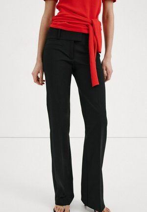 Femeie purtând pantaloni negri eleganți și un top roșu cu mâneci scurte și un nod în față, stând desculță pe un fundal alb.