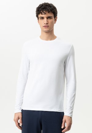 Junger Mann mit kurzen dunklen lockigen Haaren, trägt ein schlichtes weißes Langarmshirt und dunkle Hose, steht vor weißem Hintergrund.