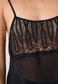Czarny top z koronką, z misternymi szczegółami szydełkowymi, cienkimi ramiączkami i przezroczystą dzianiną, podkreślający nowoczesny i stylowy design.