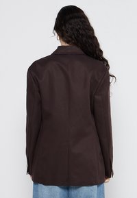 Femme aux longs cheveux bouclés foncés portant un blazer marron foncé et un jean bleu clair, vue de dos sur un fond blanc.